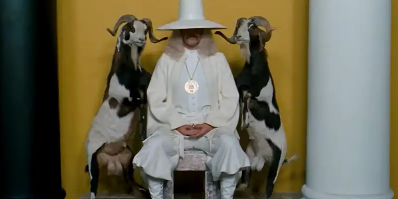 A scene from The Holy Mountain showing a man in a white suit sitting with a goat either side of him
